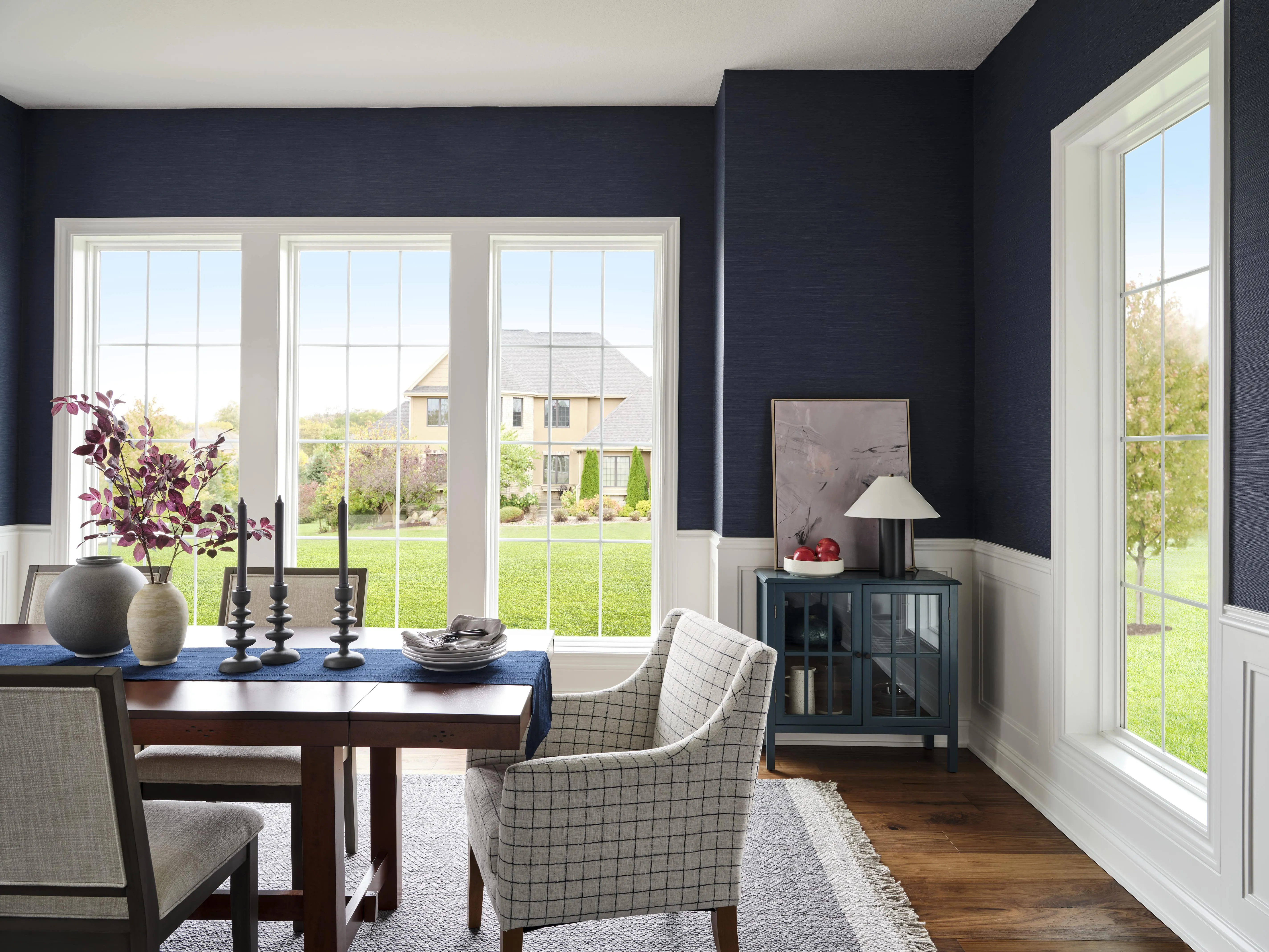 Dining room with dark blue walls, large windows overlooking a green lawn, a wooden table with gray chairs, decorative vases, black candlesticks, and a side cabinet with a lamp and artwork.